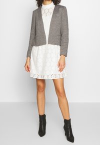 Blazer gris cropped sur une robe blanche en dentelle. Le blazer présente une texture lisse et deux poches à l'avant. Des bottines noires viennent compléter la tenue.