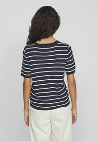 Osoba s dlouhými kudrnatými vlasy, oblečená v krátkém rukávu s námořnickými modro-bílými vodorovnými pruhy a bílými kalhotami, viděná zezadu.