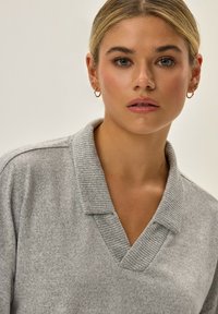 Jasnoszary sweter z prążkowanym kołnierzem i dekoltem w kształcie litery V, wykonany z teksturowanej tkaniny i o luźnym kroju. Brak zauważalnych wzorów.