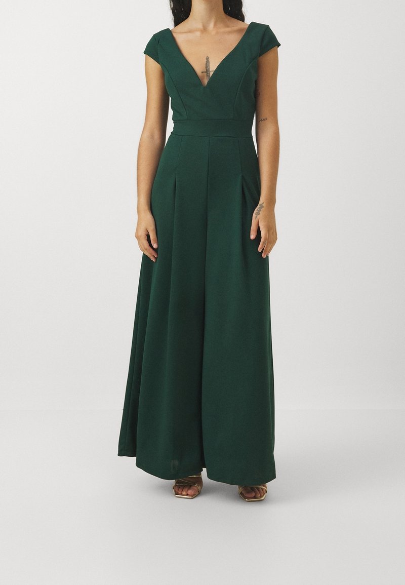 Femme portant une robe longue vert foncé avec des manches courtes et un décolleté en V profond, debout devant un fond clair uni.