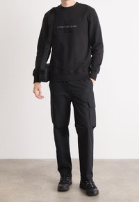 Czarna bluza z kapturem z logo "Calvin Klein Jeans", zestawiona z czarnymi spodniami cargo z bocznymi kieszeniami i czarnymi trampkami. Gładka faktura.