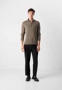 Maglione verde oliva chiaro con zip e maniche lunghe, polsini e orlo a costine, abbinato a pantaloni neri e mocassini neri. Dettaglio logo rosso.