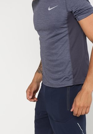 Mężczyzna w dopasowanej niebieskiej sportowej koszulce Nike i granatowych spodniach sportowych, pokazujący tatuaż na lewym przedramieniu na jednolitym tle.
