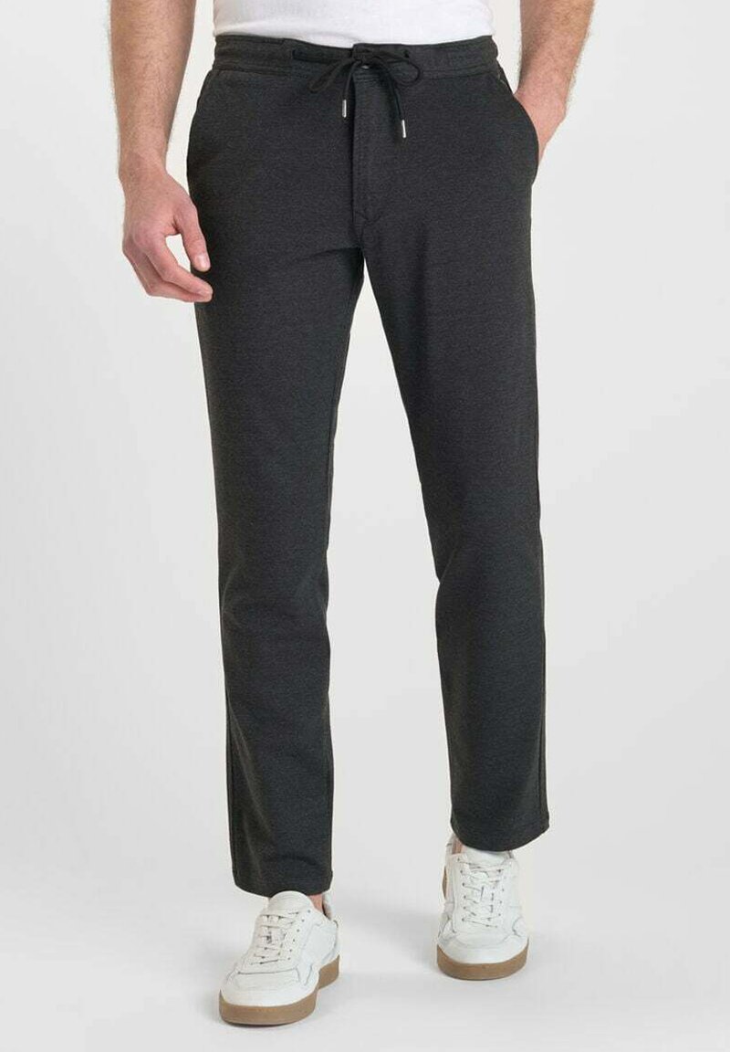 Man in een donkergrijze broek met koord en witte sneakers, staande met één hand in zijn zak tegen een effen achtergrond.