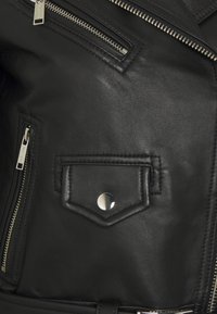 Gros plan d'une veste en cuir noire montrant des fermetures éclair argentées, une poche à rabat pressionnée et une surface en cuir texturée.