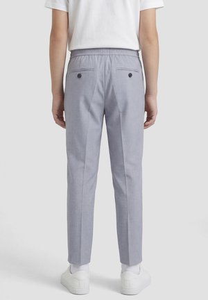 Persona che indossa pantaloni su misura grigio chiaro con tasche posteriori con bottoni, calzini bianchi, scarpe da ginnastica bianche e una camicia bianca a maniche corte, in piedi con le spalle rivolte.