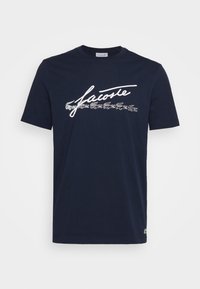 Navy blue cotton t-shirt with short sleeves, featuring white cursive "Lacoste" script and small crocodile logo near the hem.