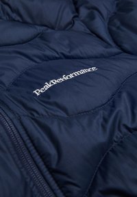 Marineblauwe pufferjack met gewatteerde textuur. Voorzien van een geborduurd logo "PeakPerformance" in het wit en een ritssluiting aan de zijkant.