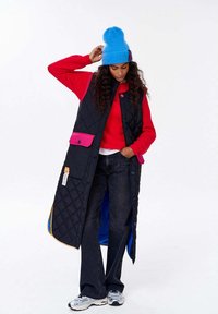 Žena v modré čepici, červeném svetru, dlouhém černém prošívaném vestě s růžovou kapsou, širokých černých džínách a bílých teniskách, stojící na bílém pozadí.