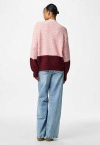 Chunky džemper u ružičastoj i burgundijskoj boji s opuštenim krojem, dugim rukavima i okruglim izrezom. Kombiniran s širokim plavim trapericama.