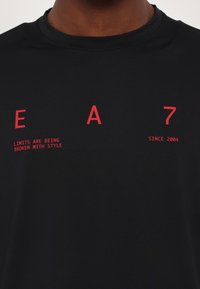 Black short-sleeve t-shirt in smooth fabric featuring red printed text: "EA 7 LIMITS ARE BEING BROKEN WITH STYLE SINCE 2004."
