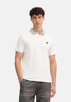 Camisa polo blanca con un cuello negro y blanco con patrón, mangas cortas y un logotipo triangular en el lado izquierdo del pecho, combinada con denim gris.