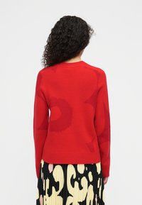 Κόκκινο πλεκτό πουλόβερ με τονικούς λουλουδένιους σχεδιασμούς και στρογγυλή λαιμόκοψη. Μαλακή υφή, μακριά μανίκια, συνδυασμένο με μια πλισέ μαύρη και κίτρινη φούστα.