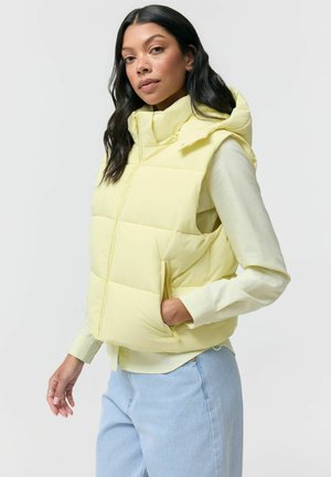 Kobieta z długimi ciemnymi włosami, ubrana w jasnosłonecznikową puchową kamizelkę na żółtej koszuli z długim rękawem oraz jasnoniebieskie dżinsy, pozująca na białym tle.