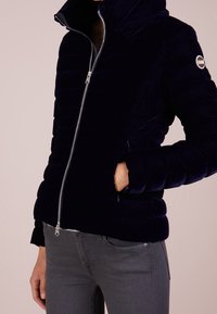 Marinblå pufferjacka med hög krage, dragkedjeslutning och ribbade muddar. Har en slät yta och logotyppatch på överarmen.