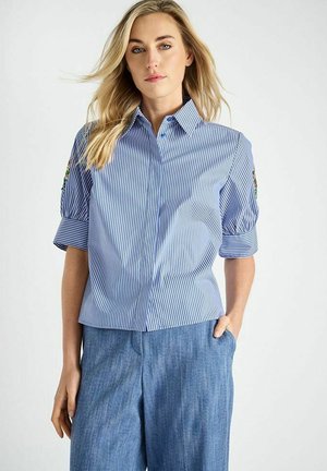 Blonde vrouw die een blauw-wit blouse met verticale strepen en bloemenborduursels op de mouwen draagt, met loszittende blauwe broek, staand met hand in de zak.