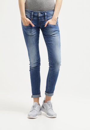 Kvinde iført opsmøgede, falmede, blå skinny jeans og grå sneakers, hænderne i lommerne, står på en hvid baggrund.