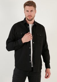 Camicia nera abbottonata in tessuto misto cotone, con due tasche sul petto, maniche lunghe e vestibilità regolare, indossata sopra una T-shirt bianca.