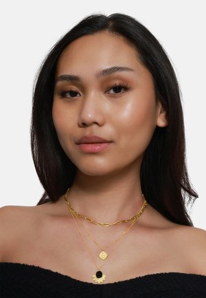 Goldfarbene, geschichtete Halsketten mit einer kräftigen Kette, einem kreisförmigen Anhänger mit schwarzem Stein und einem floralen Akzent mit filigranen Details.