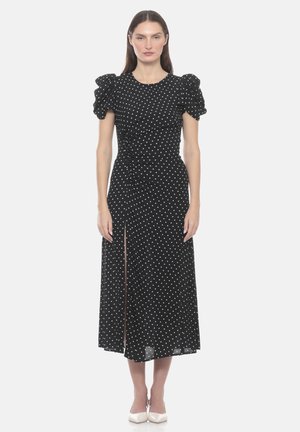 Donna che indossa un vestito midi nero con pois bianchi, maniche corte a sbuffo, spacco frontale e dettaglio arricciato, abbinato a décolleté bianchi con punta.