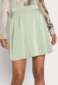 Kvinna klädd i en ljusgrön satäng minikjol och en långärmad, mönstrad nättröja, som visar låren och med händerna vilande vid sidorna.