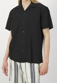 Osoba w czarnej koszuli z krótkim rękawem z guzikami i białych szortach w pionowe czarne paski, stojąca z rękami w luźnej pozycji.