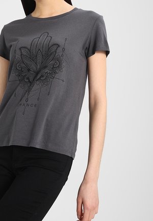 Vrouw draagt een donkergrijs T-shirt met korte mouwen, met een zwart ingewikkeld blad- en abstract patroon en het woord "CHANGE" op de voorkant.