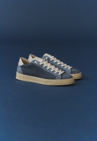 Paire de baskets basses bleu marine et beige avec lacets blancs sur une surface et un fond bleu foncé.