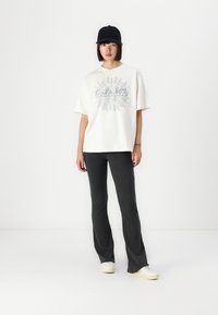 T-shirt bianca in cotone oversize con un design grafico argentato, abbinata a pantaloni grigi scuri a zampa d'elefante e sneakers bianche, completata da un cappellino nero.