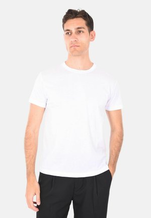 Giovane uomo con capelli scuri che indossa una semplice maglietta bianca a maniche corte e pantaloni neri, in piedi con una mano in tasca.