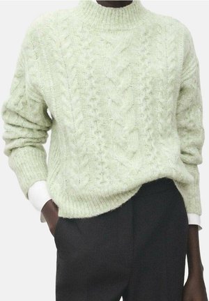 Pull vert clair à torsades porté sur une chemise blanche, associé à un pantalon taille haute foncé, avec une main dans la poche.