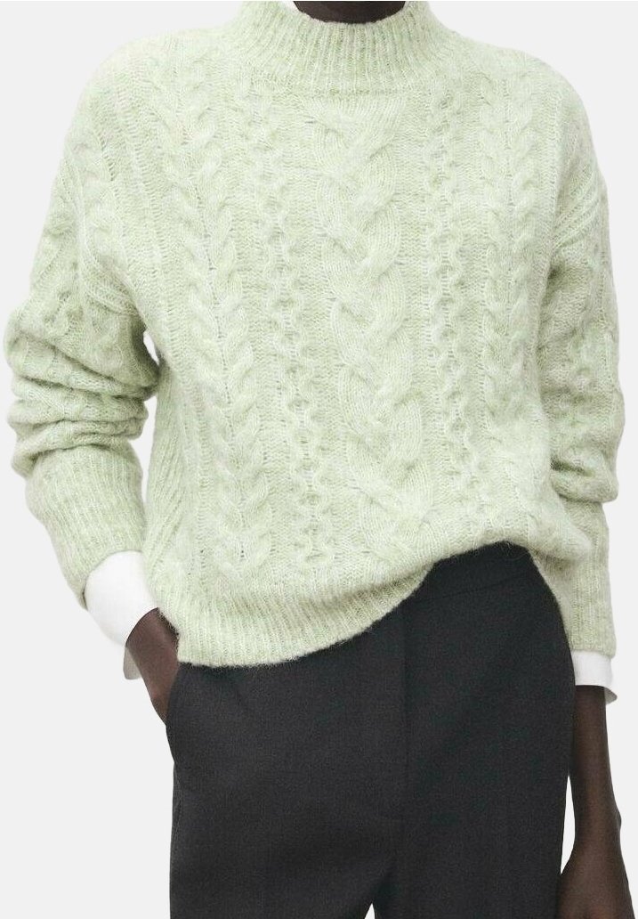 Pull vert clair à torsades porté sur une chemise blanche, associé à un pantalon taille haute foncé, avec une main dans la poche.