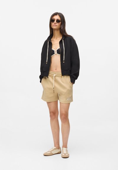 Veste courte noire avec cordons de serrage, short beige et lunettes de soleil noires. Le haut de bikini est en tissu noir avec un détail de fermeture blanc.