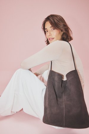 Bolso tote de ante gris con un diseño minimalista. Presenta una amplia apertura y una textura suave. Modelo vestido con una blusa clara y pantalones blancos, sentado/a contra un fondo rosa.