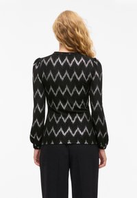 Mujer con cabello rubio largo y ondulado lleva una blusa negra de mangas largas con un patrón en zigzag plateado y pantalones negros, de pie frente a un fondo blanco.