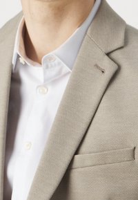 Uomo che indossa un blazer beige texturizzato sopra una camicia bianca a maniche lunghe, con colletto e bottoni visibili, mostrato dalla nuca fino a metà torso.