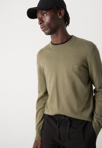 Maglione a maglia verde oliva con colletto e polsini a coste, dotato di una piccola toppa con logo beige. Abbinato a pantaloni neri con coulisse.