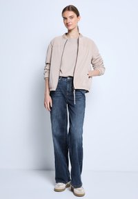 Bomber jacket beige claro con acabado suave, top beige claro, jeans rectos azul oscuro y zapatillas de color claro.