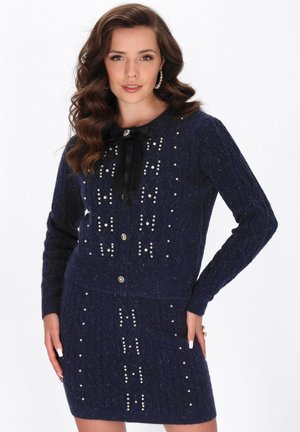 Vrouw met golvend bruin haar draagt een marineblauwe gebreide cardigan en rokset, versierd met parels en een zwarte strik bij de kraag.