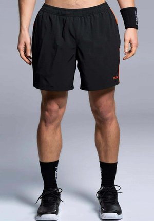 Homme portant un short de sport noir, des chaussettes noires avec lettres blanches, des chaussures de sport noires et un bracelet noir, debout, jambes légèrement écartées.