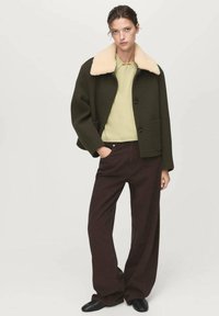 Vrouw in een donker olijfgroene jas met een beige schapenhuiden kraag, lichte groene trui, brede bruine broek en zwarte schoenen, staande met één hand in de zak.