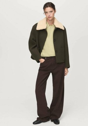 Donna con una giacca verde scuro con colletto in shearling beige, maglione verde chiaro, pantaloni larghi marroni e scarpe nere, in piedi con una mano in tasca.