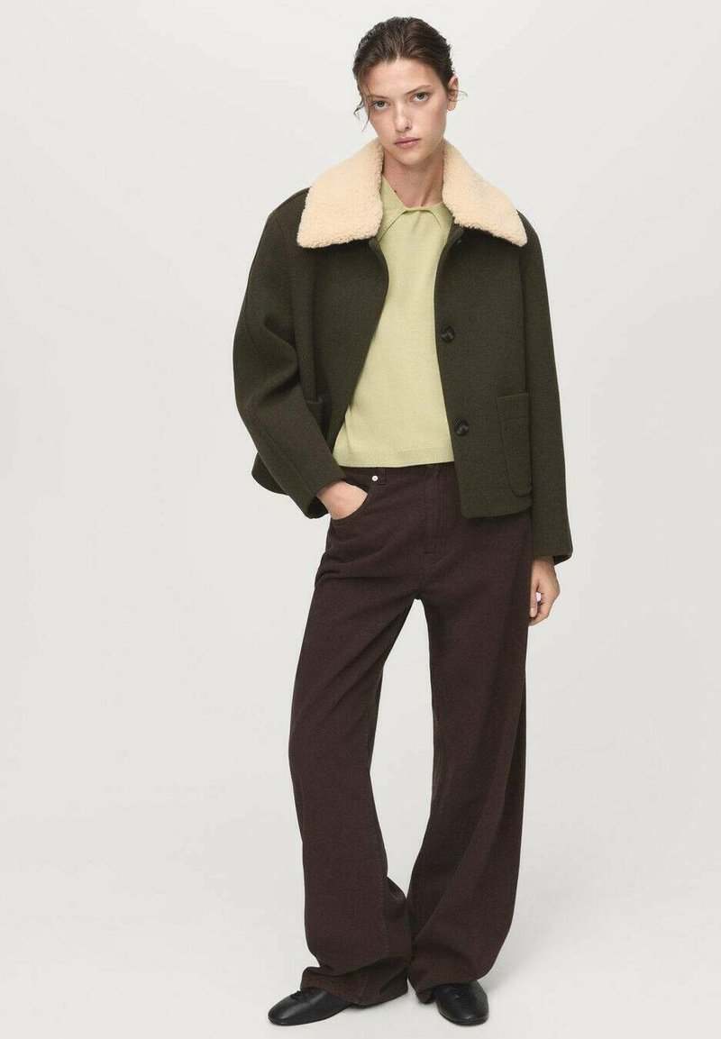 Vrouw in een donker olijfgroene jas met een beige schapenhuiden kraag, lichte groene trui, brede bruine broek en zwarte schoenen, staande met één hand in de zak.