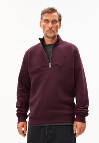 Bordeaux sweatshirt met ritssluiting, gemaakt van zachte stof, met een horizontale naad en ribbelmanchetten. Gekozen boven een zwart t-shirt.