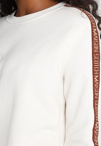 Felpa bianca con colletto a costine e tessuto testurizzato. Presenta una striscia marrone a contrasto con logo lungo il lato.