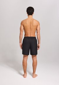 Schwarze Badehose mit elastischem Bund, knie-langer Gestaltung und glatter Textur, von hinten an einem nackten männlichen Modell gezeigt.