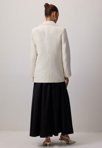 Biely textúrovaný sako s dlhými rukávmi, viditeľný detail na chrbte, kombinované s voľnou čiernou sukňou a béžovými podpätkami. Minimalistický design, elegantná silueta.
