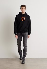 Sudadera con capucha negra y un logo naranja brillante, combinada con jeans grises y botas de cuero negras, contra un fondo liso.