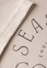 Tejido de algodón beige con una textura suave, que presenta texto impreso en oscuro en una tipografía en negrita. La costura está perfectamente cosida a lo largo del borde.