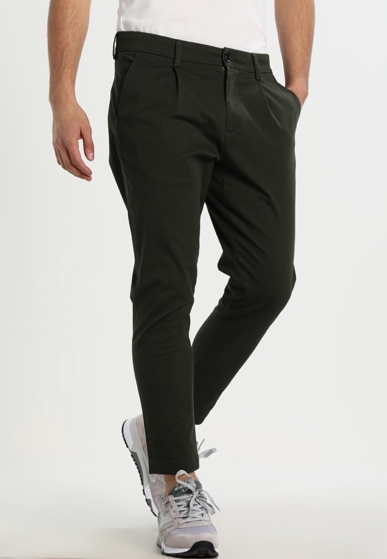 Homme portant un pantalon noir slim avec poches latérales, des baskets blanches et une chemise blanche, debout avec une main dans la poche.